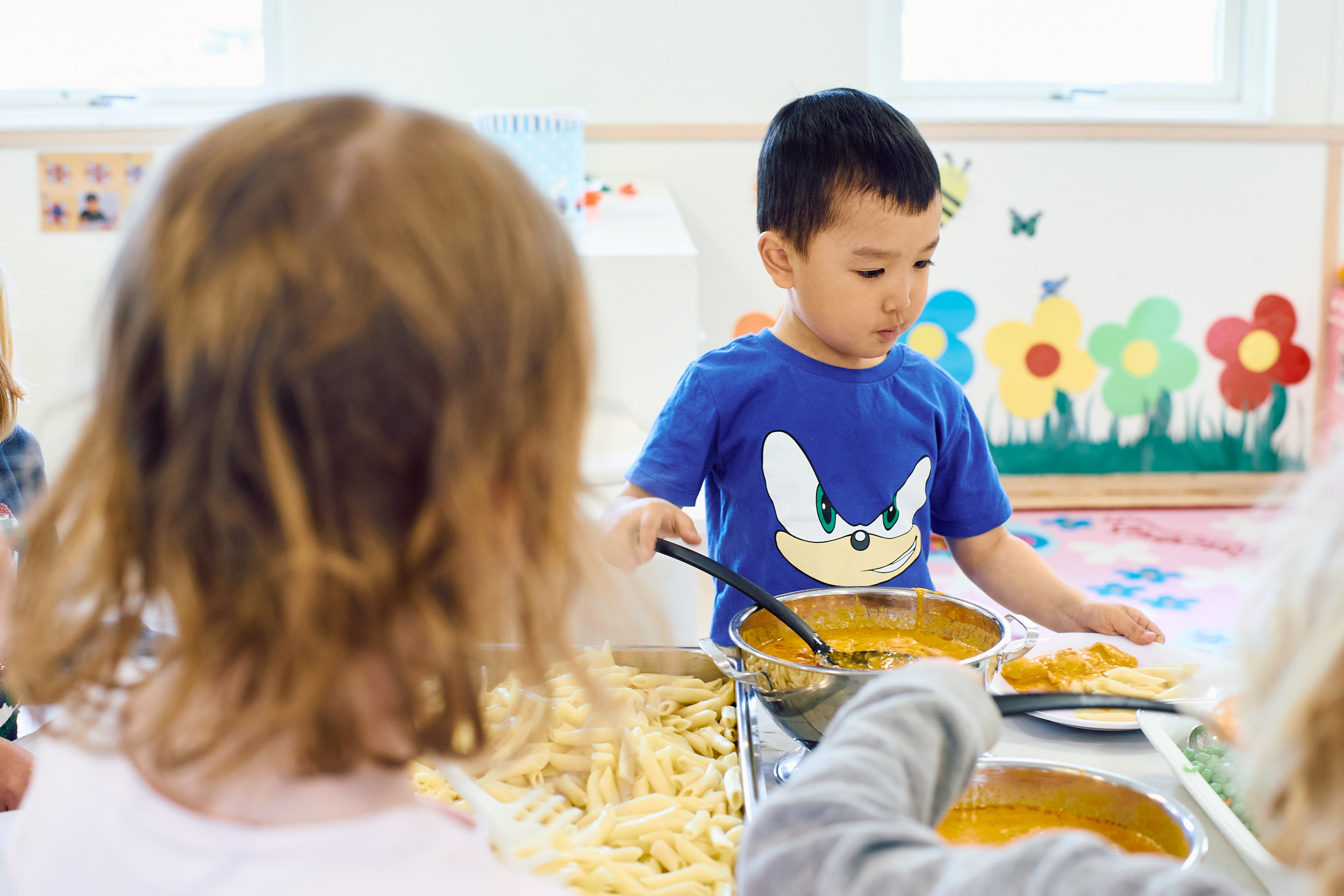 Förskolebarns bakhuvud syns i förgrunden. Längre in står en pojke i blå tröja och tar mat från en skål.